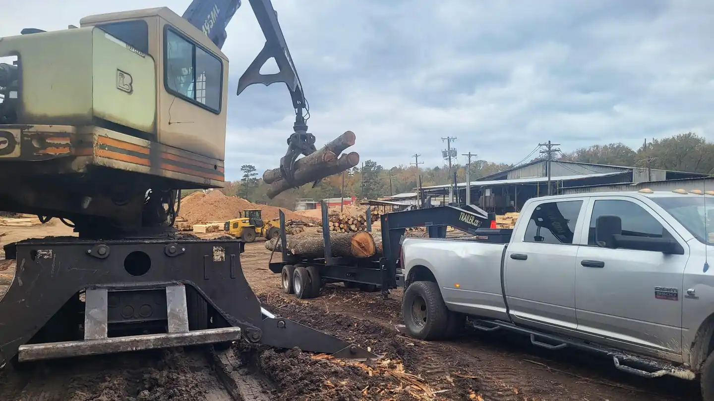 Lester's knuckle-boom grapple log loader picking up a whole log beside a pickup and a log trailer