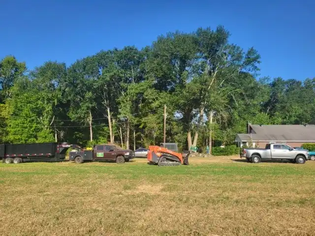 Dump trailers, trucks and an orange track loader lined up in a field below tall trees