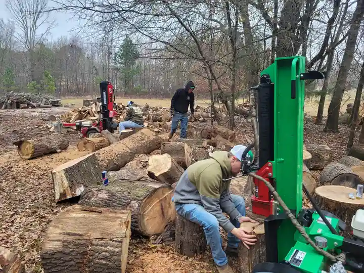 Lester's crew splitting cut rounds with a green hydraulic splitter in a wooded yard full of logs