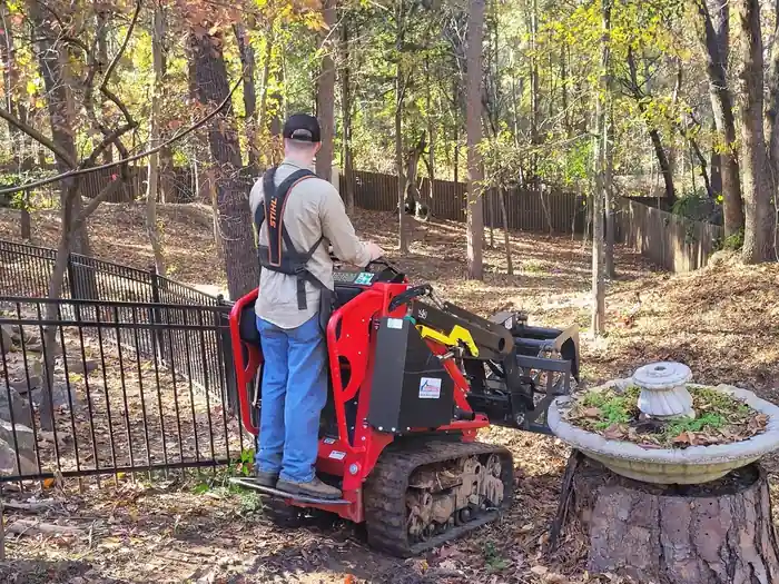 An operator riding a red stand-on mini loader beside a wrought-iron fence and a birdbath, autumn leaves on the ground