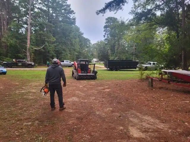 A crew member with a chainsaw on a red-dirt lot, a track loader with a mulcher behind him